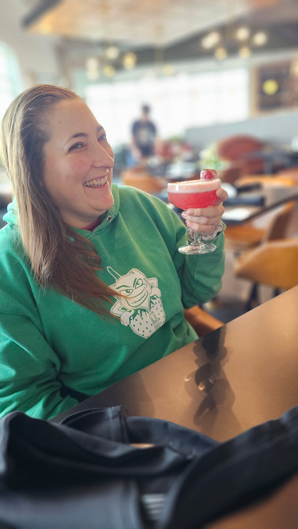 Krista in a green Fired Up sweatshirt with a logo, drinking a mocktail with our Strawberry Vanilla Rhubarb Jam used in it