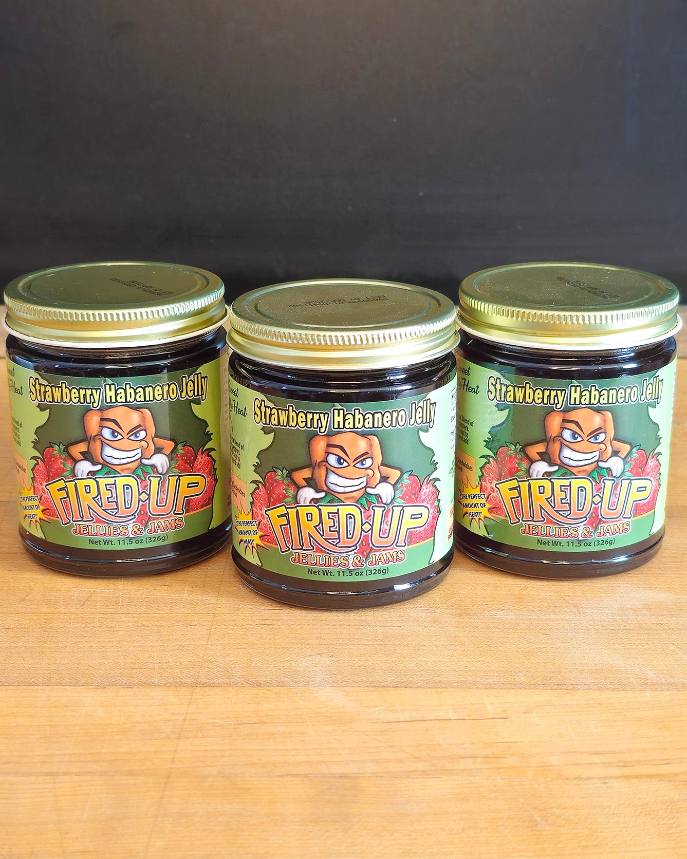 Three jars of Strawberry Habanero Jelly 11.5 oz labeled 'Fired Up Jellies and Jams' on a wooden surface with a dark background.