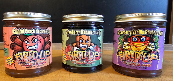 Three jars of Fired Up jellies and jams, each labeled with different flavors: Cinnful Peach Habanero, Strawberry Habanero, and Strawberry Vanilla Rhubarb, arranged side by side on a cutting board