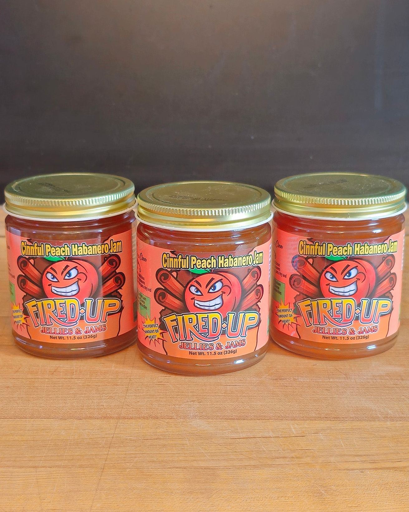Three jars of 'Fired Up Jellies and Jams' Cinnful Peach Habanero Jam on a wooden surface with a dark background