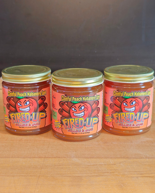 Three jars of 'Fired Up Jellies and Jams' Cinnful Peach Habanero Jam on a wooden surface with a dark background