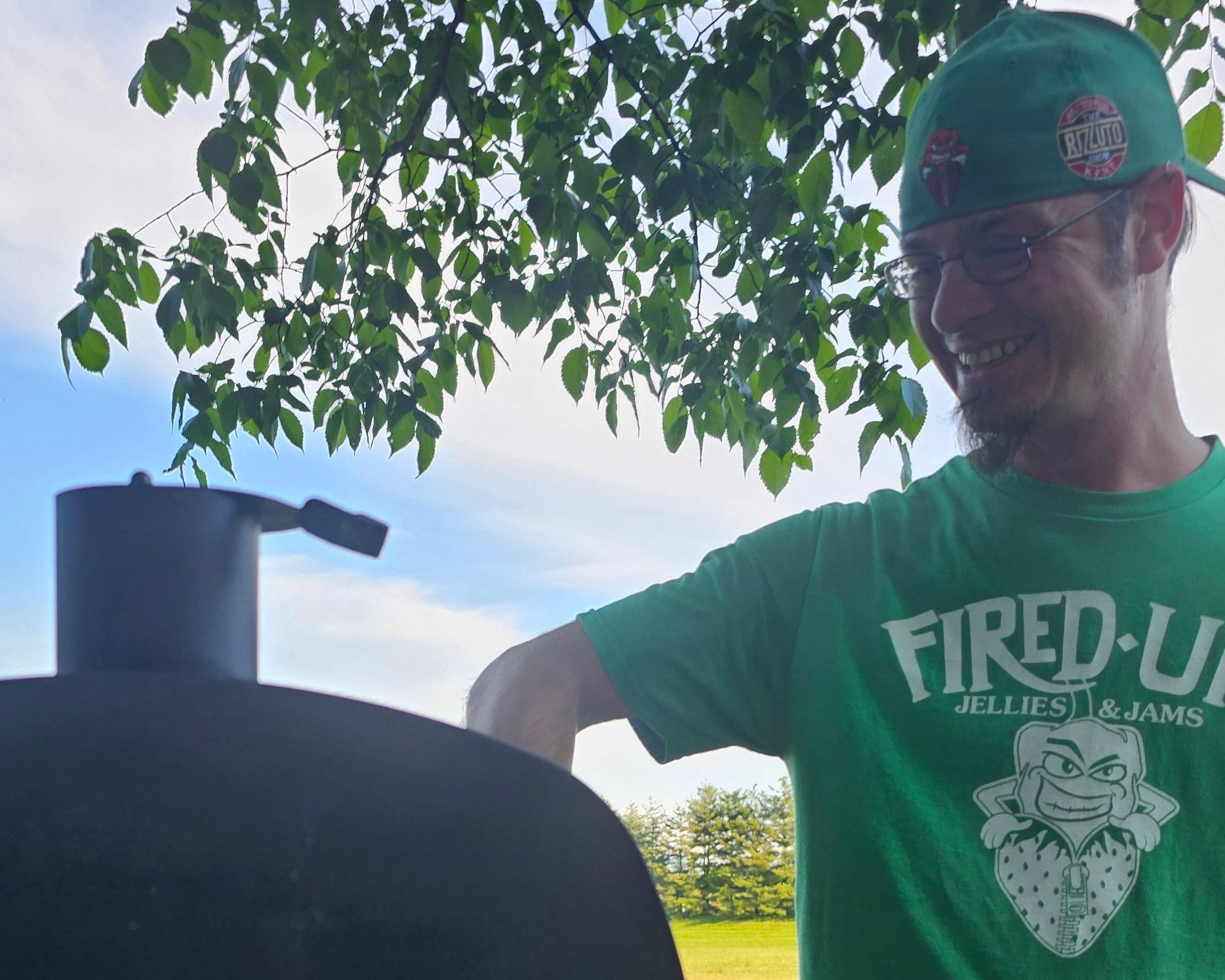 Smitty wearing a green Fired Up t-shirt, standing outdoors next to a barbecue grill.